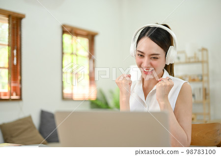 Cheerful Asian woman wearing headphones, looking at laptop screen, celebrating her success. 98783100
