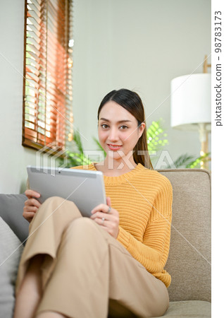 Beautiful Asian woman using her tablet on sofa, smiling and looking at the camera. 98783173