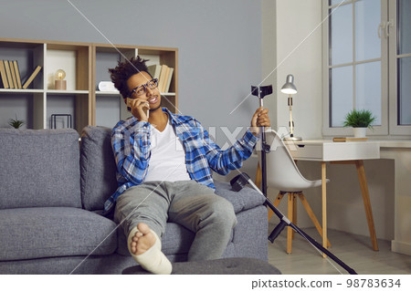 Handsome young man with fractured leg sitting on sofa talking on cell phone 98783634