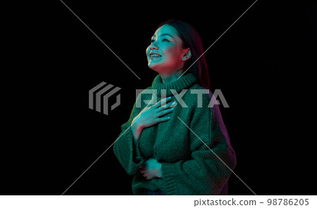 Very excited. Young attractive brunette girl in cozy sweater looking away with interest over dark background in neon light. Concept of emotions, youth lifestyle Very excited. Young attractive brunette girl in cozy sweater looking away with interest over dark background in neon light. Concept of emotions, youth lifestyle 98786205