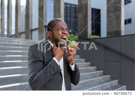 An African-American businessman is standing on the street near an office center and blowing an aerosol into his throat. He feels severe pain in the throat, cough, allergy, suffocation. An African-American businessman is standing on the street near an office center and blowing an aerosol into his throat. He feels severe pain in the throat, cough, allergy, suffocation. 98786673