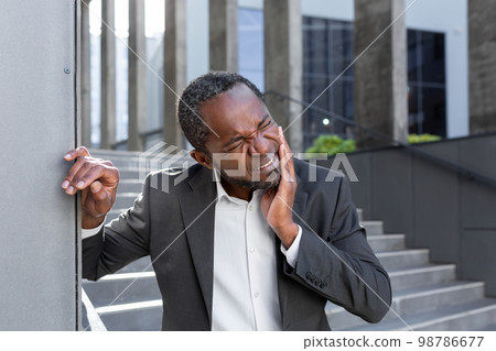 Toothache outside. An African-American man in a suit is standing near an office center, holding his cheek, leaning against a wall. He feels a strong toothache, discomfort in his mouth. 98786677