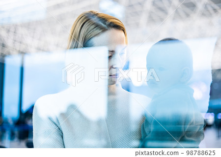 Thoughtful young mother looking trough window holding his infant baby boy child while waiting to board an airplane at airport terminal departure gates. Travel with baby concept 98788625
