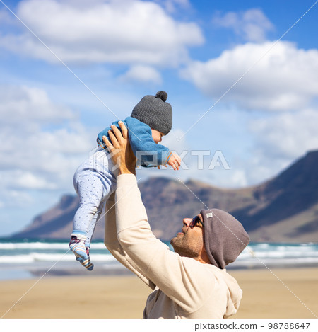 Father enjoying pure nature holding and playing with his infant baby boy sun in on windy sandy beach of Famara, Lanzarote island, Spain. Family travel and parenting concept 98788647