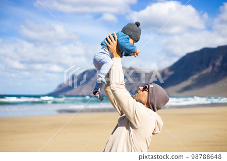 Father enjoying pure nature holding and playing with his infant baby boy sun in on windy sandy beach of Famara, Lanzarote island, Spain. Family travel and parenting concept 98788648