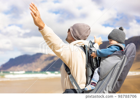 Young father rising hands to the sky while enjoying pure nature carrying his infant baby boy sun in backpack on windy sandy beach of Famara, Lanzarote island, Spain. Family travel concept Young father rising hands to the sky while enjoying pure nature carrying his infant baby boy sun in backpack on windy sandy beach of Famara, Lanzarote island, Spain. Family travel concept 98788706
