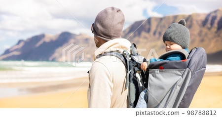 Young father carrying his infant baby boy sun in backpack on windy sandy beach of Famara, Lanzarote island, Spain. Family travel and winter vacation concept. Parenting, fatherhood, father's day 98788812
