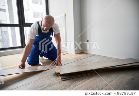 Man in work overalls installing laminate wood flooring in new flat. Male construction worker laying laminate wooden planks on floor underlayment. Home renovation concept. Man in work overalls installing laminate wood flooring in new flat. Male construction worker laying laminate wooden planks on floor underlayment. Home renovation concept. 98789187