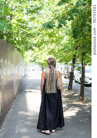A girl in a black dress and long afro-braids walks along a summer street, rear view A girl in a black dress and long afro-braids walks along a summer street, rear view 98791081