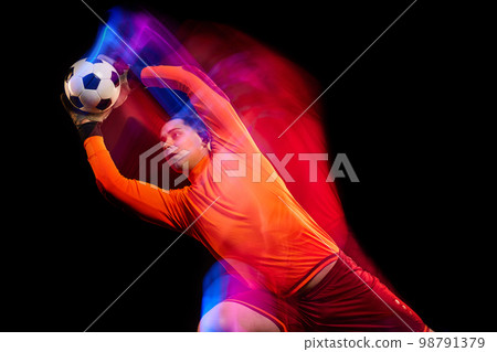 Sportive energetic man, soccer football goalkeeper in action, motion over dark background with mixed neon light. Concept of sport, achievement, competition, goals. Sportive energetic man, soccer football goalkeeper in action, motion over dark background with mixed neon light. Concept of sport, achievement, competition, goals. 98791379