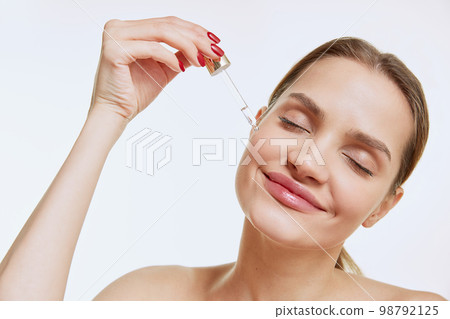 Portrait of young beautiful girl with clear face applying face serum, taking care after skin condition over grey studio background. Concept of natural beauty, cosmetology Portrait of young beautiful girl with clear face applying face serum, taking care after skin condition over grey studio background. Concept of natural beauty, cosmetology 98792125