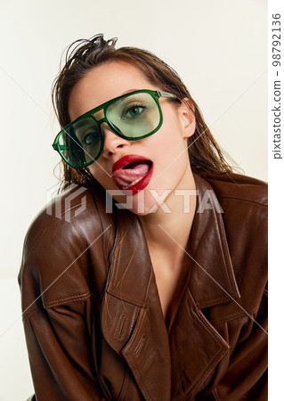 Sensual femininity. Beautiful young brunette girl in stylish leather jacket and green trendy sunglasses posing over grey studio background. Concept of beauty, fashion Sensual femininity. Beautiful young brunette girl in stylish leather jacket and green trendy sunglasses posing over grey studio background. Concept of beauty, fashion 98792136