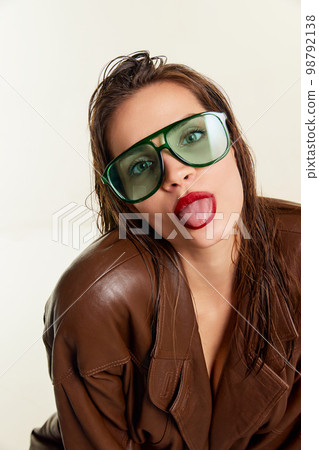 Sticking out tongue grimace. Beautiful young brunette girl in stylish leather jacket and green sunglasses posing over grey studio background. Concept of beauty, fashion 98792138