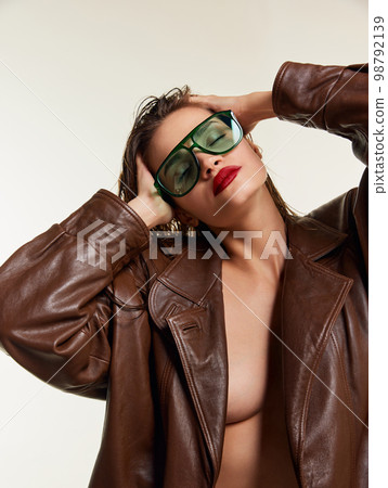 Attractive sensual portrait of beautiful young brunette girl with wet hair in stylish leather jacket, green sunglasses posing over grey studio background. Concept of beauty, fashion Attractive sensual portrait of beautiful young brunette girl with wet hair in stylish leather jacket, green sunglasses posing over grey studio background. Concept of beauty, fashion 98792139