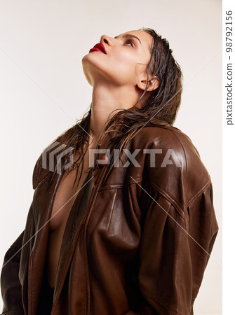 Attractive youth. Beautiful young brunette girl with wet hair and red lips in stylish leather jacket posing over grey studio background. Concept of beauty, fashion 98792156