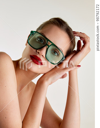 Extravagant style and femininity. Young beautiful girl with red lips posing in green sunglases over grey studio background. Concept of natural beauty, skin care, cosmetology Extravagant style and femininity. Young beautiful girl with red lips posing in green sunglases over grey studio background. Concept of natural beauty, skin care, cosmetology 98792172