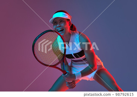 Portrait of young professional female tennis player in sports uniform in motion, action over gradient pink-purple background in neon light. Sport, fashion, energy, motivation concept Portrait of young professional female tennis player in sports uniform in motion, action over gradient pink-purple background in neon light. Sport, fashion, energy, motivation concept 98792615