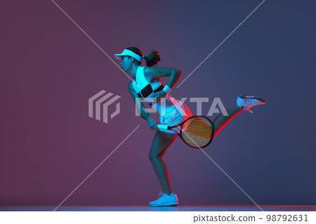 Portrait of young professional female tennis player in sports uniform in motion, action over gradient pink-purple background in neon light. Sport, fashion, energy, motivation concept 98792631