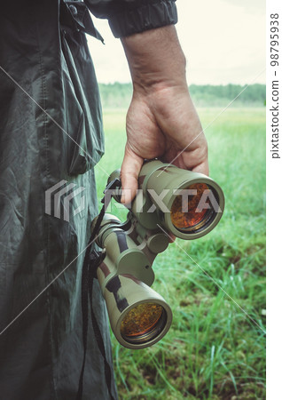 closeup view of hand of huntsman or traveller or border guardian in dark khaki raincoat holding green binoculars with red lens against background of grassy field in forest or swamp closeup view of hand of huntsman or traveller or border guardian in dark khaki raincoat holding green binoculars with red lens against background of grassy field in forest or swamp 98795938
