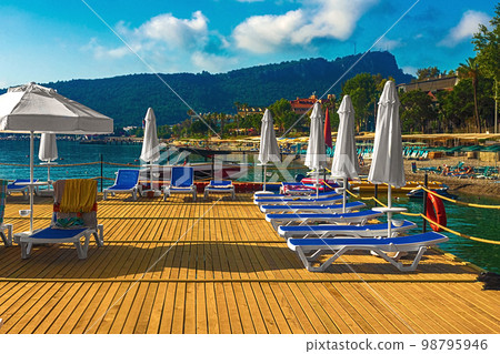 View of the pier with blue sun loungers and white umbrellas on the seashore in Kemer. Turkey View of the pier with blue sun loungers and white umbrellas on the seashore in Kemer. Turkey 98795946