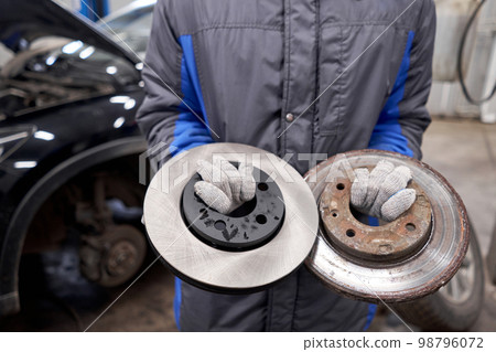 The mechanic holds old rusty brake disc and new disc. Change the old to new brake disc on car in a garage. Auto repair concept. The mechanic holds old rusty brake disc and new disc. Change the old to new brake disc on car in a garage. Auto repair concept. 98796072