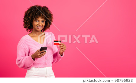 Cheerful young black curly lady shopaholic in casual with braces uses smartphone, credit card to check financial account 98796928