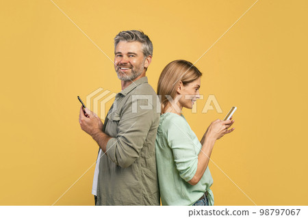Modern lifestyle. Happy middle aged couple standing back to back, using their cellphones, man smiling at camera 98797067