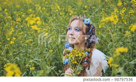 A young girl poses in a rapeseed field with a beautiful hairdo of flowers and butterflies. 98797536