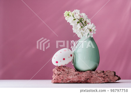 Happy Easter. Easte egg and blooming white lilac branches in small vase on a rock against magenta background. copy space 98797758