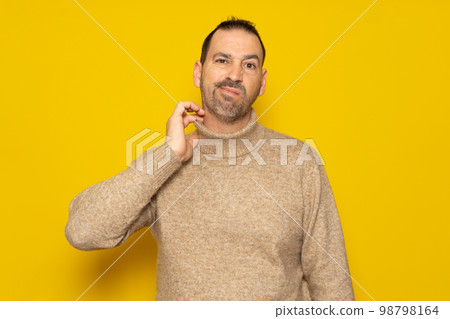 Bearded hispanic man wearing beige turtleneck scratching his neck and grimacing isolated over yellow background. Concept of doubt and indecision. 98798164