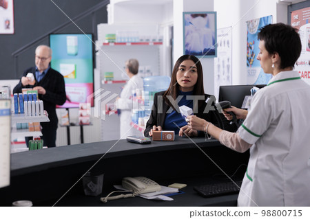 Young asian woman buying sunscreen and waiting at pharmacy checkout. Pharmaceutical worker wearing medical retail store uniform scanning medicaments packages bar code at counter desk Young asian woman buying sunscreen and waiting at pharmacy checkout. Pharmaceutical worker wearing medical retail store uniform scanning medicaments packages bar code at counter desk 98800715