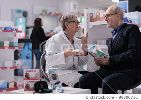 Drugstore employee explaining heart supplements dosage instruction to patient. Elderly man listening to pharmacy assistant talking about vitamin benefit and side effects in apothecary Drugstore employee explaining heart supplements dosage instruction to patient. Elderly man listening to pharmacy assistant talking about vitamin benefit and side effects in apothecary 98800716