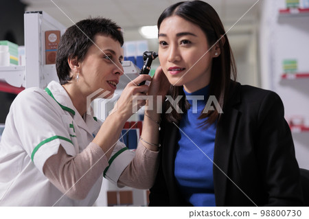 Pharmacist looking in patient ear and discussing infection treatment in pharmacy store. Pharmaceutical assistant using otoscope medical tool for disease diagnostics in drugstore Pharmacist looking in patient ear and discussing infection treatment in pharmacy store. Pharmaceutical assistant using otoscope medical tool for disease diagnostics in drugstore 98800730