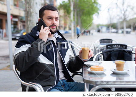 Young man talking on phone during breakfast in street cafe Young man talking on phone during breakfast in street cafe 98802700