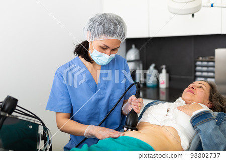 Focused young female cosmetologist in a protective mask makes an elderly woman client a vacuum massage of the abdomen Focused young female cosmetologist in a protective mask makes an elderly woman client a vacuum massage of the abdomen 98802737