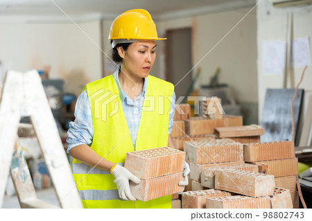 Workwoman in yellow vest and hardhat carrying hollow clay bricks on construction site indoors 98802743
