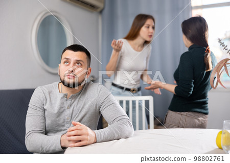 Sad man sitting at table at home while two women quarreling Sad man sitting at table at home while two women quarreling 98802761
