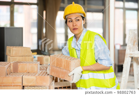 Workwoman in yellow vest and hardhat carrying hollow clay bricks on construction site indoors Workwoman in yellow vest and hardhat carrying hollow clay bricks on construction site indoors 98802763