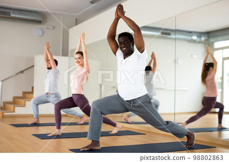 Man exercising warrior one yoga pose with people in gym Man exercising warrior one yoga pose with people in gym 98802863