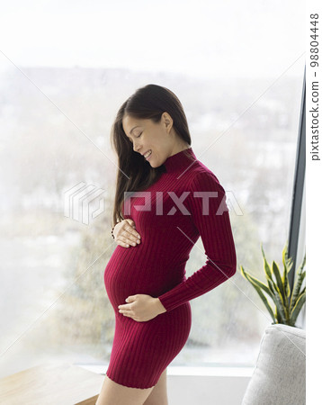 Pregnant woman at home. Prenatal pregnancy photo of beautiful Asian model standing holding belly in red dress indoors with winter background. 2nd trimester 98804448