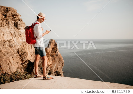 Digital nomad, man in the hat, a businessman with a laptop sits on the rocks by the sea during sunset, makes a business transaction online from a distance. Remote work on vacation. 98811729