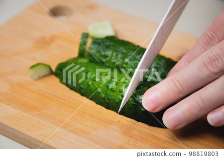 Female fingers with a knife cut a cucumber, close-up, on a wooden cutting board 98819803