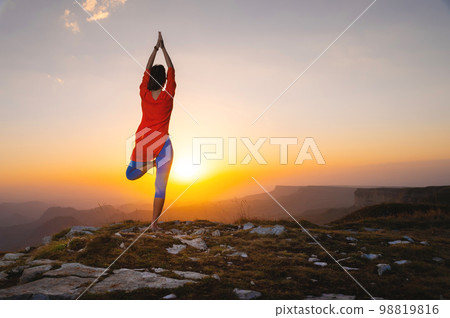 Full length rear view of a woman standing on one leg while doing yoga in the mountains, shot at sunset during summer vacation 98819816