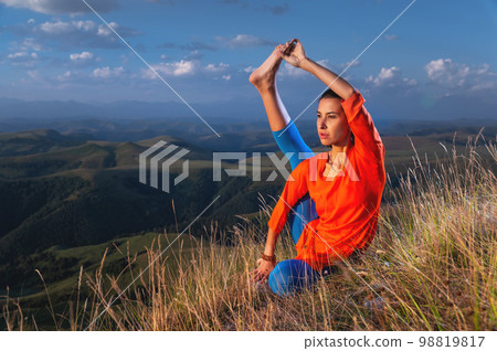 Young attractive yogi woman practicing yoga, doing variation of Krounchasana exercises, Heron pose, exercising in nature, wearing sportswear, mountains 98819817