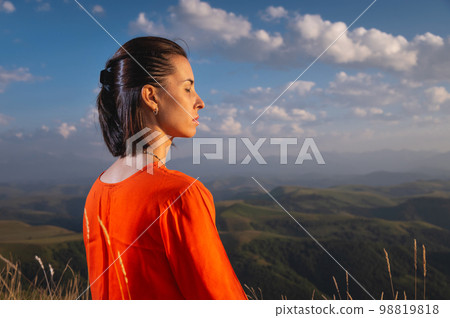Portrait of a woman with closed eyes, focused on meditation, against the backdrop of a cloudy sky in the mountains and sunlight falling on her 98819818