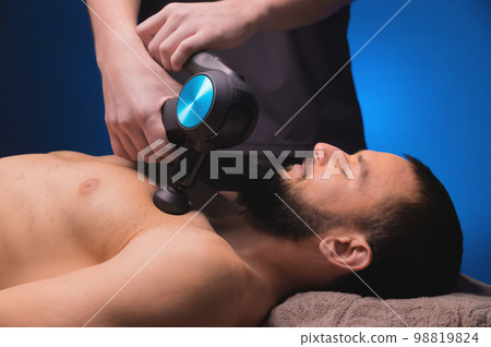 Athletic young man lies on his back, his chest is massaged with a massage gun in the salon, he warms up after training, close-up 98819824