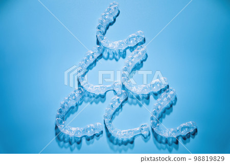 Close up, invisible aligners on a blue background in the shape of a pyramid or triangle creating a pattern. Plastic braces for teeth alignment Close up, invisible aligners on a blue background in the shape of a pyramid or triangle creating a pattern. Plastic braces for teeth alignment 98819829