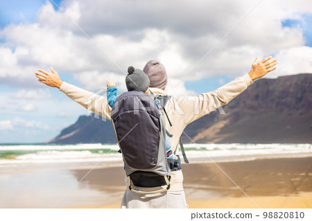 Young father rising hands to the sky while enjoying pure nature carrying his infant baby boy son in backpack on windy sandy beach of Famara, Lanzarote island, Spain. Family travel concept 98820810