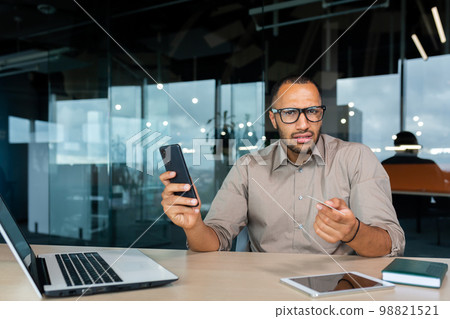 Dissatisfied businessman inside office looking at camera, hispanic man holding bank credit card and phone in hands, cheated man got rejection and error when trying to make online money transfer. 98821521