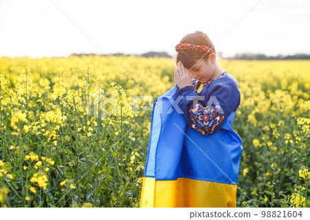 Pray for Ukraine. Child with Ukrainian flag in rapeseed field. Little girl holding national flag praying for peace. 98821604